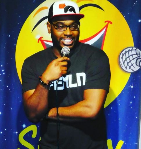 Comedian Ricarlo Winston, performing live comedy show, smiling and holding microphone.